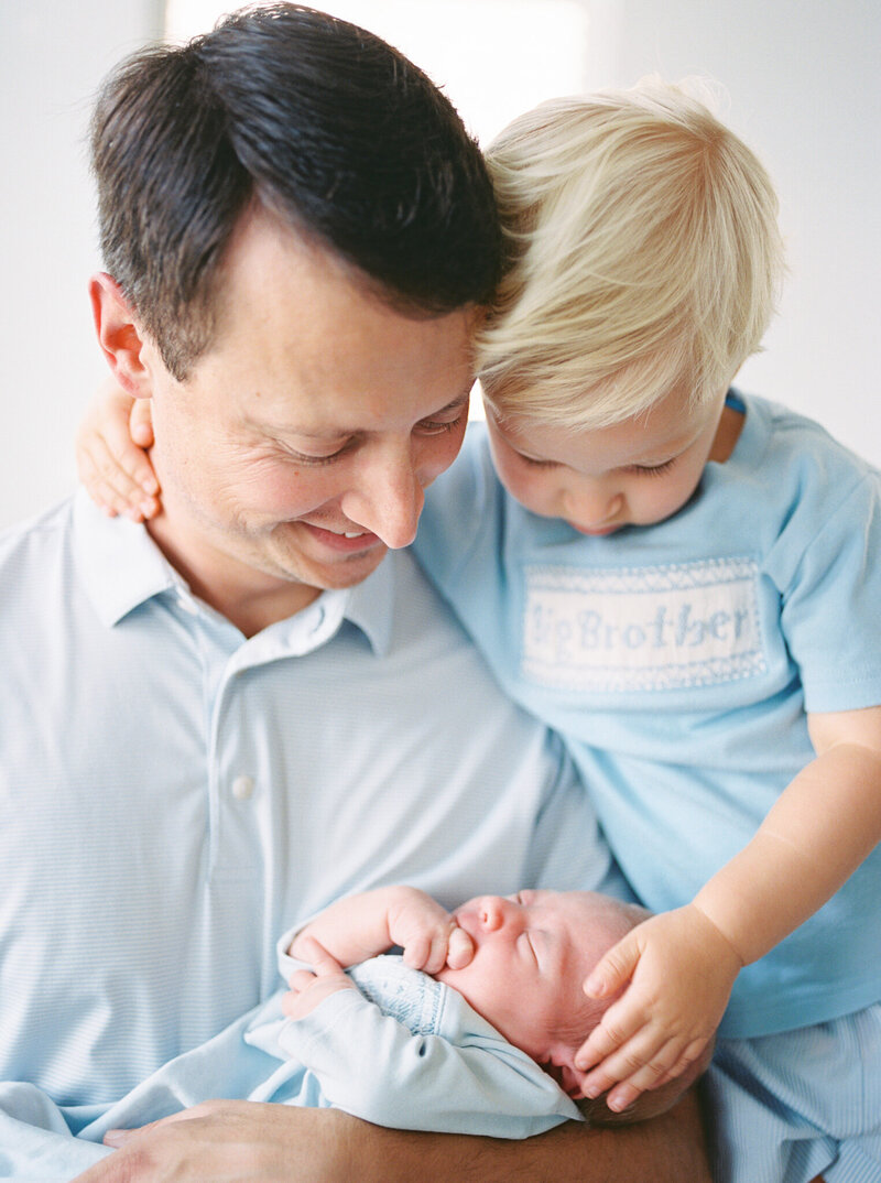 Brunette father in pale blue shirt looks down at newborn son in his arms while blonde, toddler son in blue shirt holds dad's neck and also looks down at baby