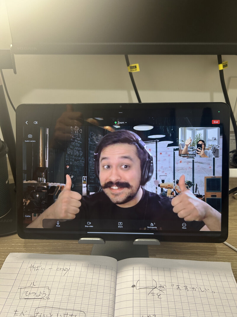 Smiling student giving two thumbs up on a video call during a Japan Blend coaching session, notebook visible on desk