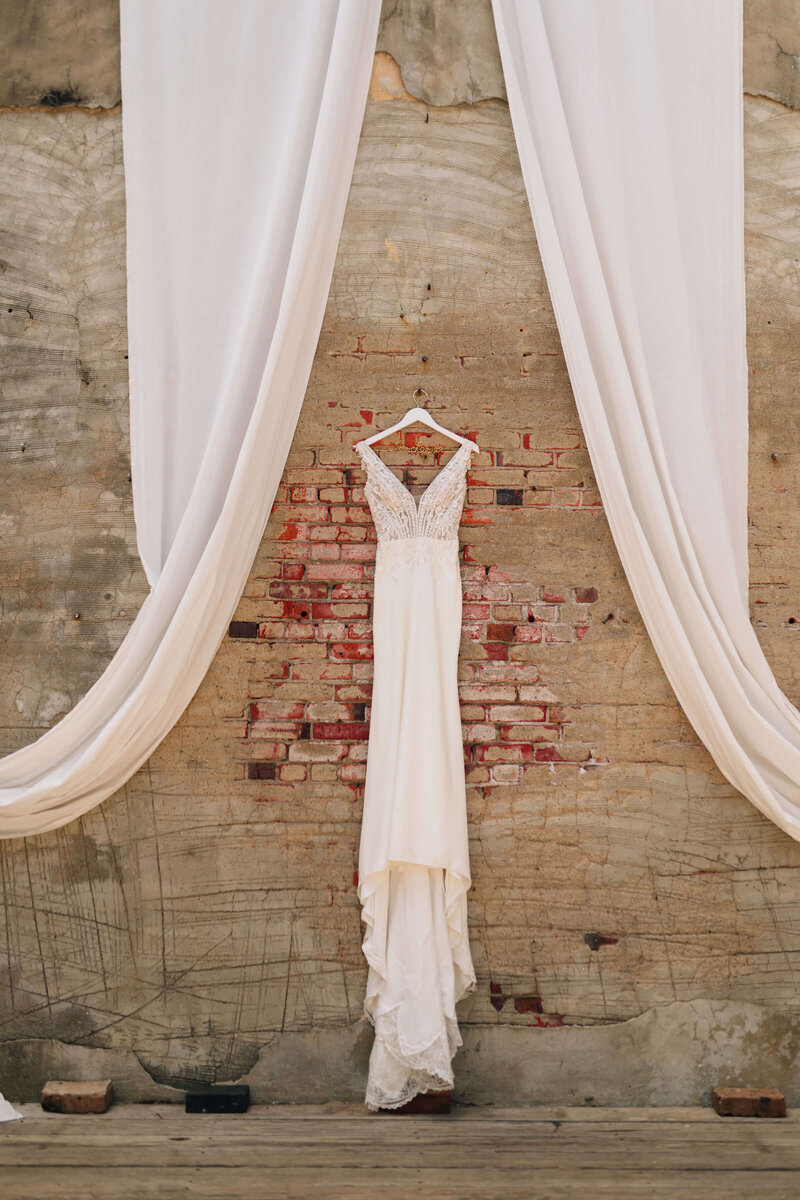 A white wedding dress hangs on a wooden hanger against a brick and concrete wall, framed by flowing white drapes on either side.