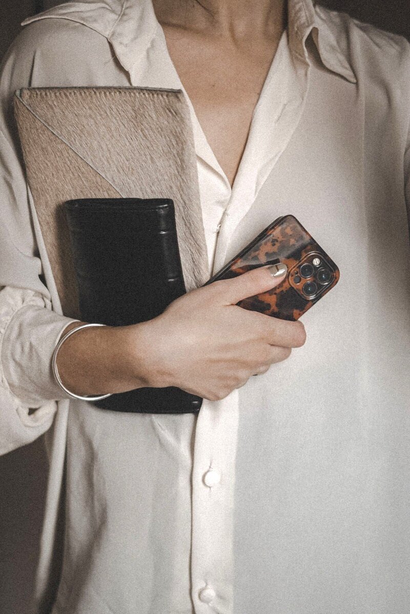 A woman, draped in an oversized cream silk blouse, holds a folio, black purse, and tortoiseshell iPhone close to her chest, her posture one of calm assurance as she stands in quiet anticipation.