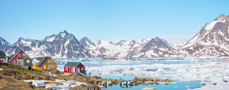 Colorful wooden houses sit along a rocky shore with snow-capped mountains and floating ice in the background under a clear blue sky.