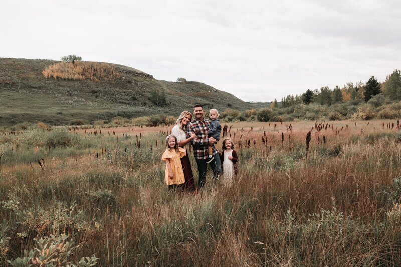 A man kisses his fiance on the cheek during their enagagement session in a Alberta foothills field. She has her eyes closed and is smiling with her hand on his chest