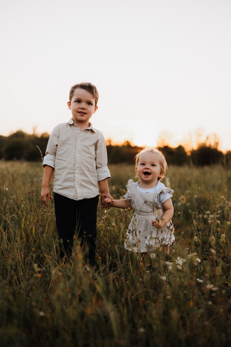 Bruder und Schwester stehen Händchen haltend auf einer Wiese während Golden Hour
