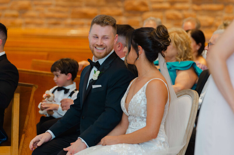 a groom in a black tuxedo smiling at his bride during their church ceremony.  Captured by Ottawa wedding photographer JEMMAN Photography