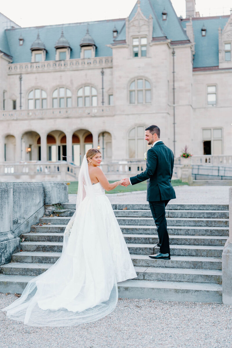 Newport Wedding Photographer | A bride and groom stand on stone steps, holding hands. The bride wears a flowing white gown, and the groom is in a dark suit. A grand building is in the background.
