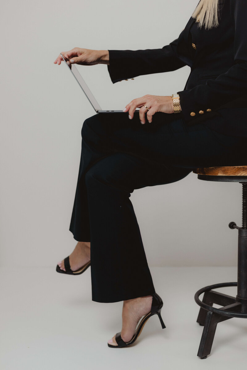 Professional woman in a black suit and heels sitting on a stool, working on a laptop—capturing a confident, modern business aesthetic.