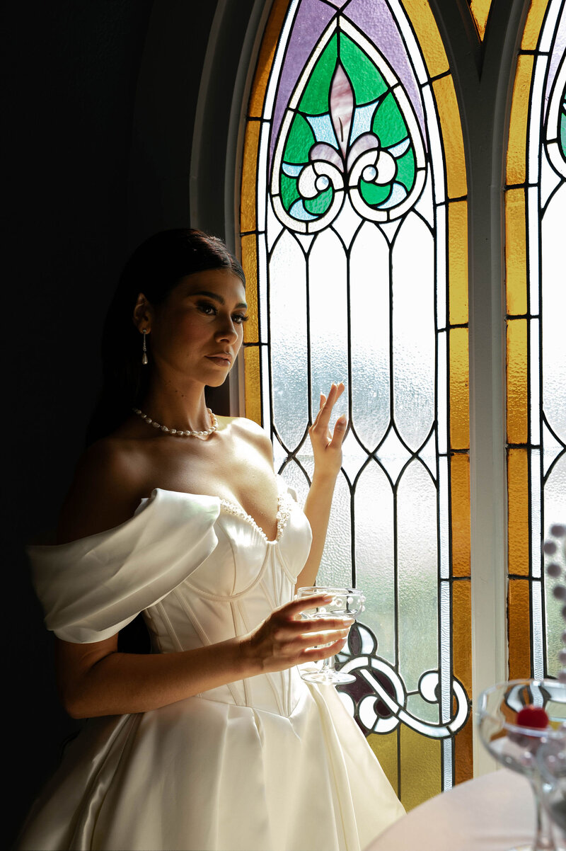 A bride in an elegant off-shoulder gown gazes thoughtfully out a vibrant stained glass window, her expression serene and reflective.