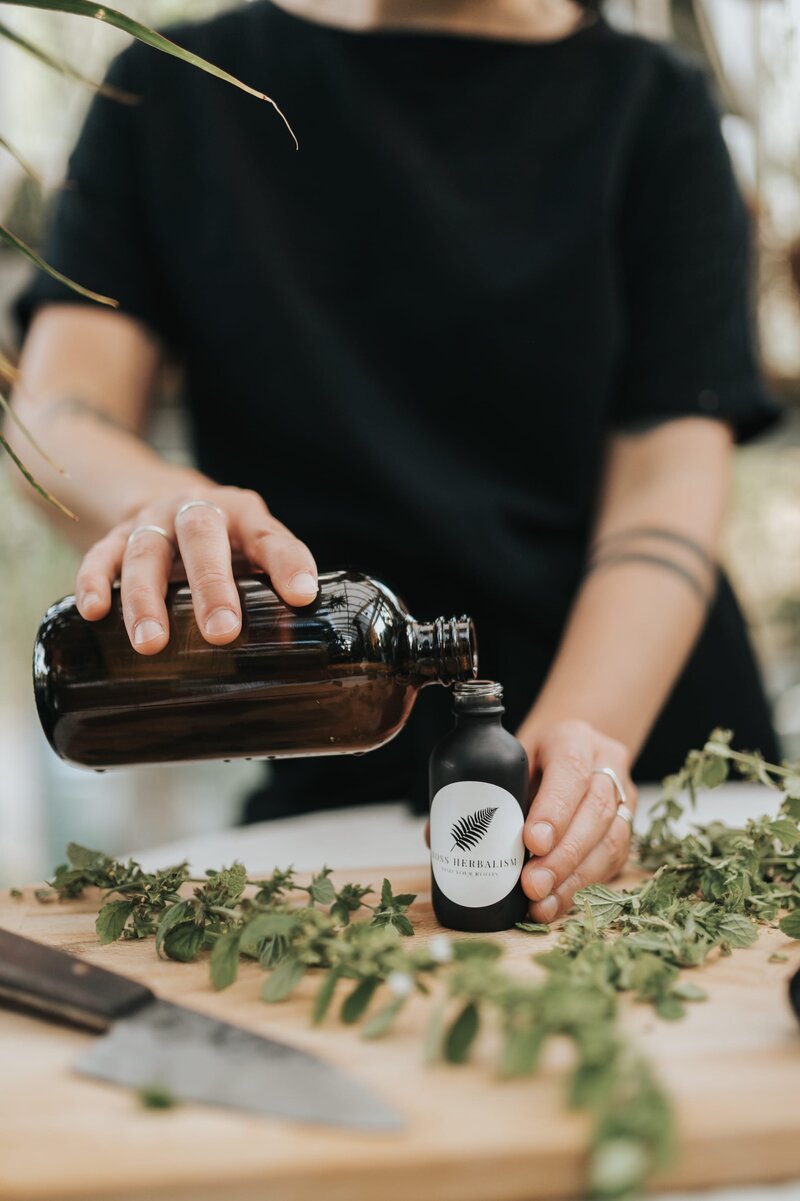 Woman wearing black shirt pours liquid from large amber bottle into small black bottle with a sticker that reads "Moss Herbalism" with a fern illustration above it,