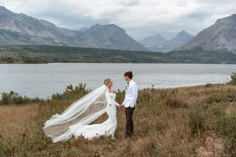 A bride and groom stand hand in hand beside St. Mary Lake in Glacier National Park as her veil flows dramatically in the wind, surrounded by rugged peaks and soft gray skies, captured by Sydney Breann Photography.