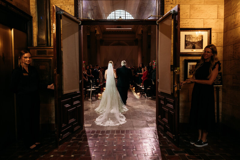 Bride and father walk down the aisle as doors open to the glowing ceremony space at Treasury on the Plaza with guests watching.