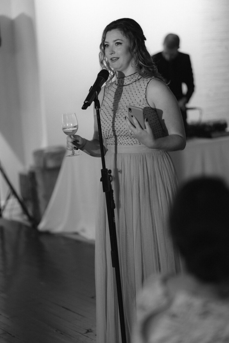 A woman delivers a speech at an event as guests seated at a table listen attentively.