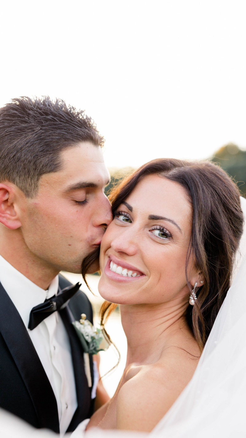 Grrom giving bride a kiss on the cheek. While she is smiling to the camera