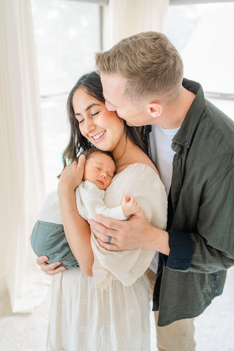 a mother holds her newborn on her chest while her husband stands behind her and kisses her on the cheek.