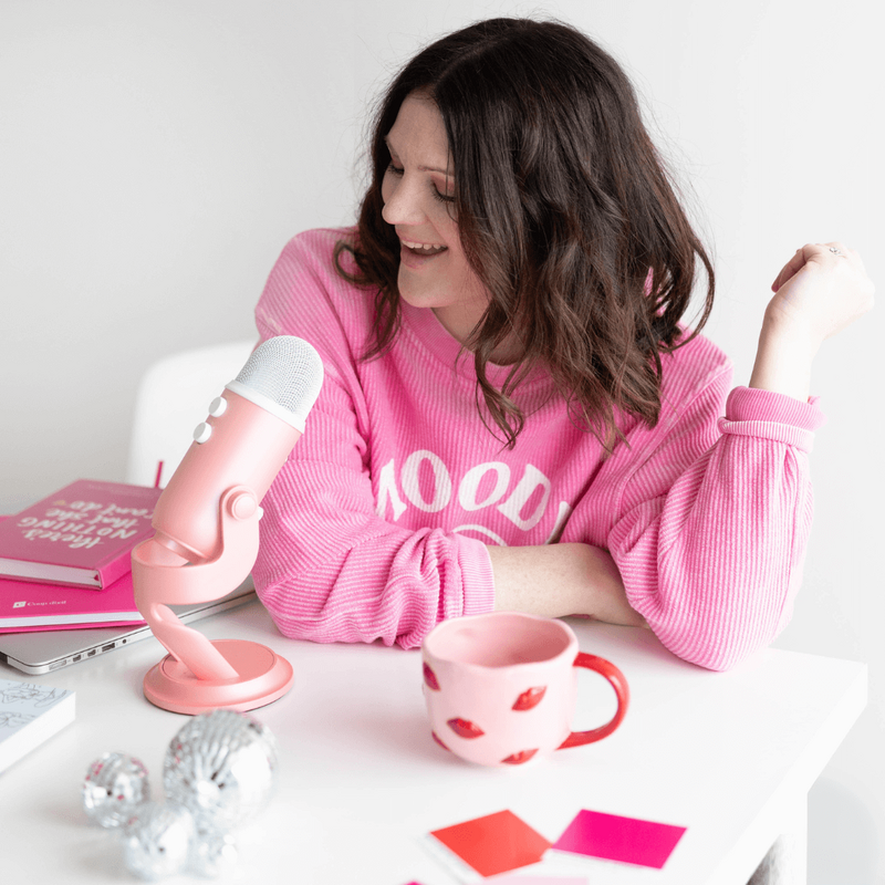 Emily, a brand and web designer, sits at her desk in a pink sweatshirt, speaking into a microphone.