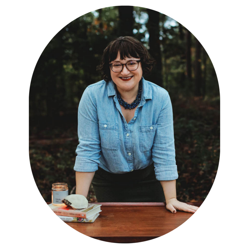 Ash Stinson smiles at the camera while leaning over a wood desk stacked with books and trees in the background