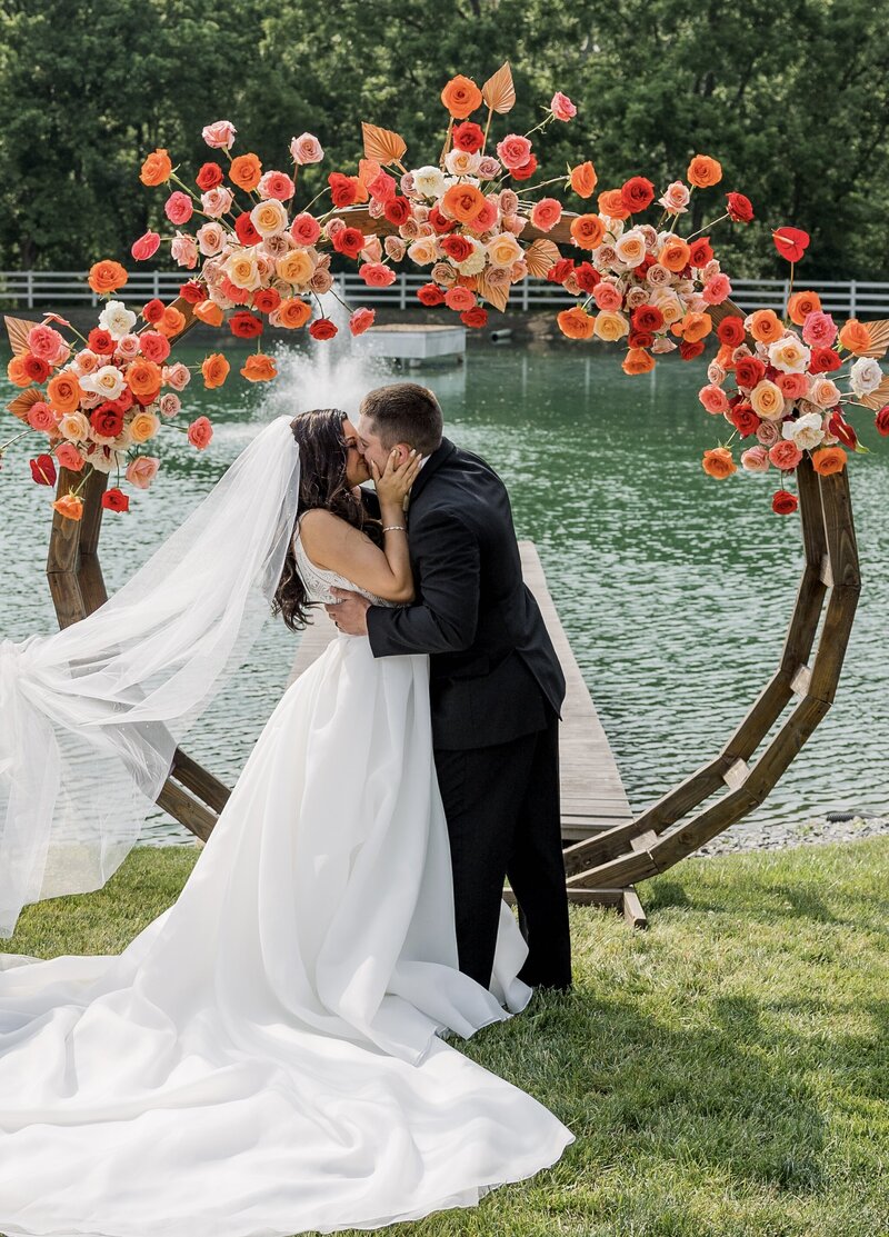 Bride and groom kissing in front of circular ceremony centerpiece decorated with bright red and orange florals