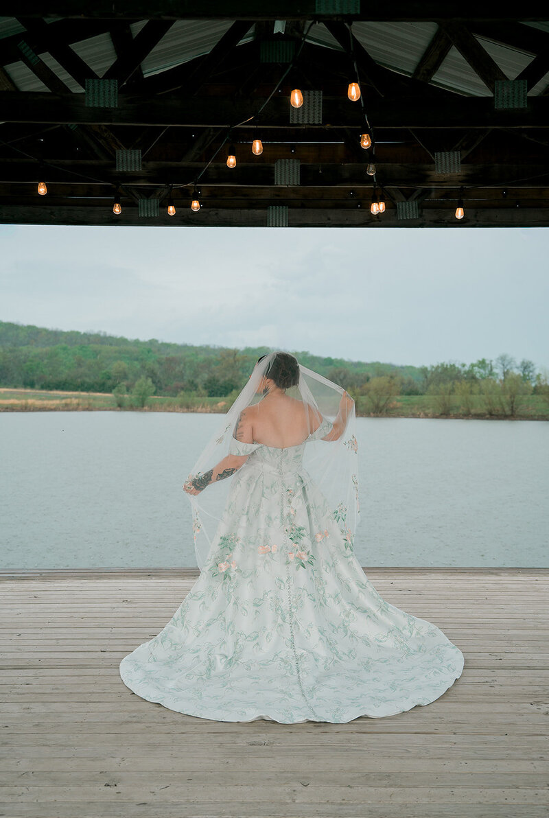 A bride stands facing a lake. She is holding her hands out to the sides inside her veil. 