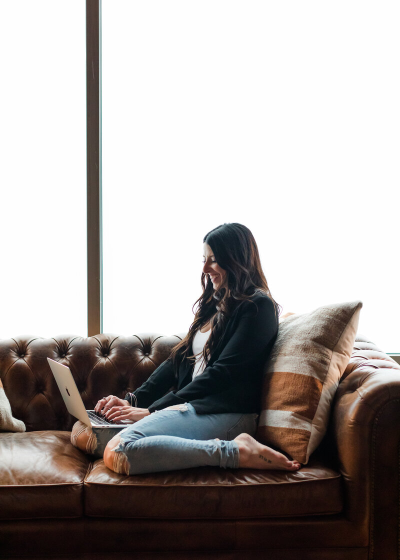 Allie Clark sits cross-legged on a brown leather couch with a striped pillow, working on her laptop in a relaxed, natural light setting.
