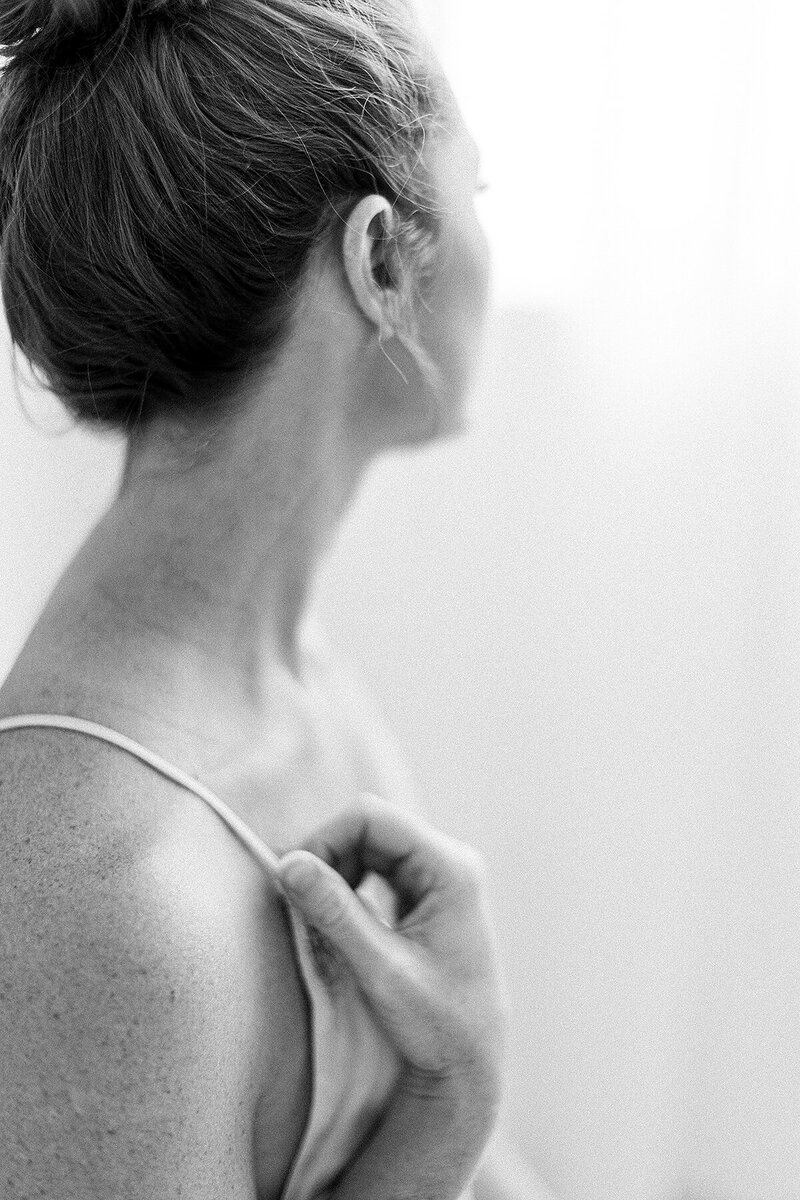 A black and white boudoir portrait of a woman delicately holding her bra strap while looking out of a window in a photography studio located in Dallas, TX