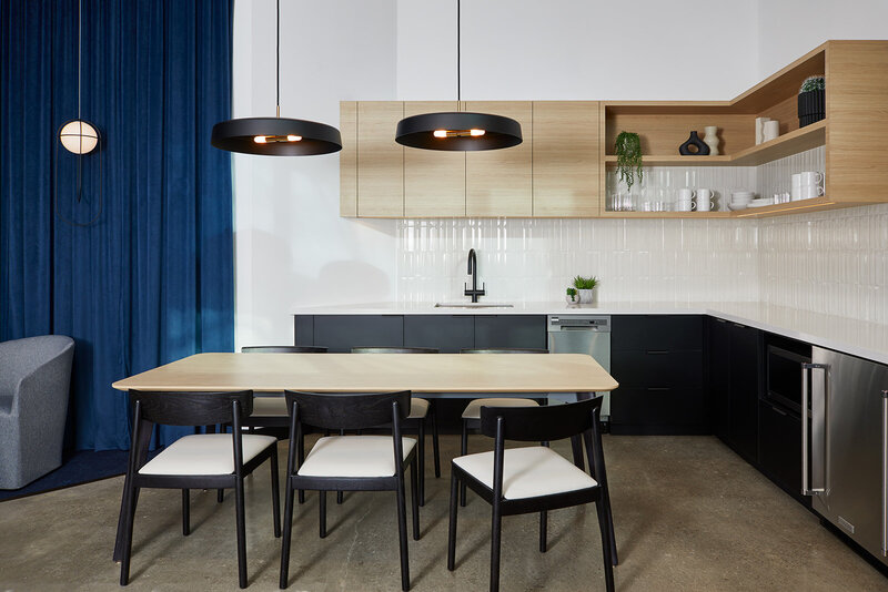 A cafe seating area containing a wood table and chairs in front of a blue curtain backdrop and small kitchenette featuring cabinetry with sleek black and wood finishes