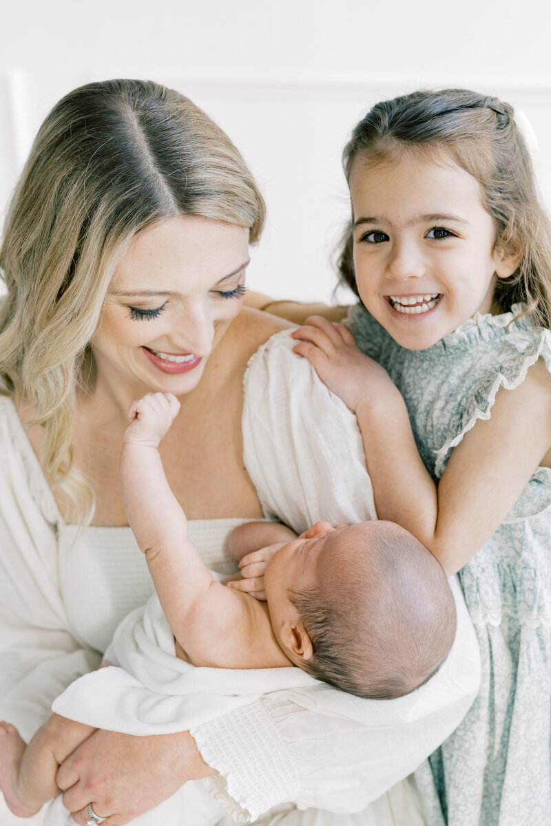 Mother gently holding newborn with big sister smiling nearby during lifestyle motherhood session, Charlotte newborn photographer