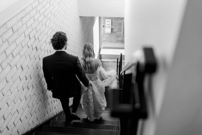 groom holds brides dress train while walking down the stairs