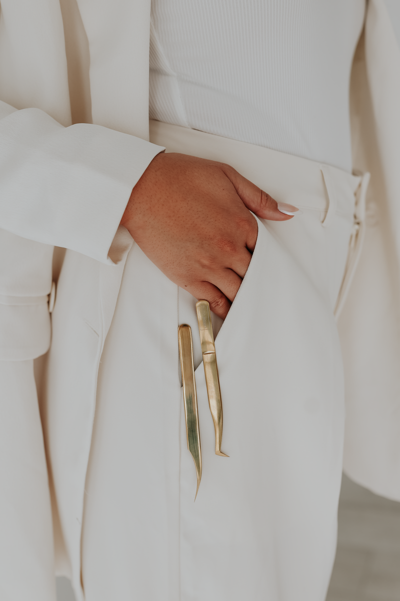 woman in white suit with hands in pockets along with a pair of eyelash extension tweezers. 