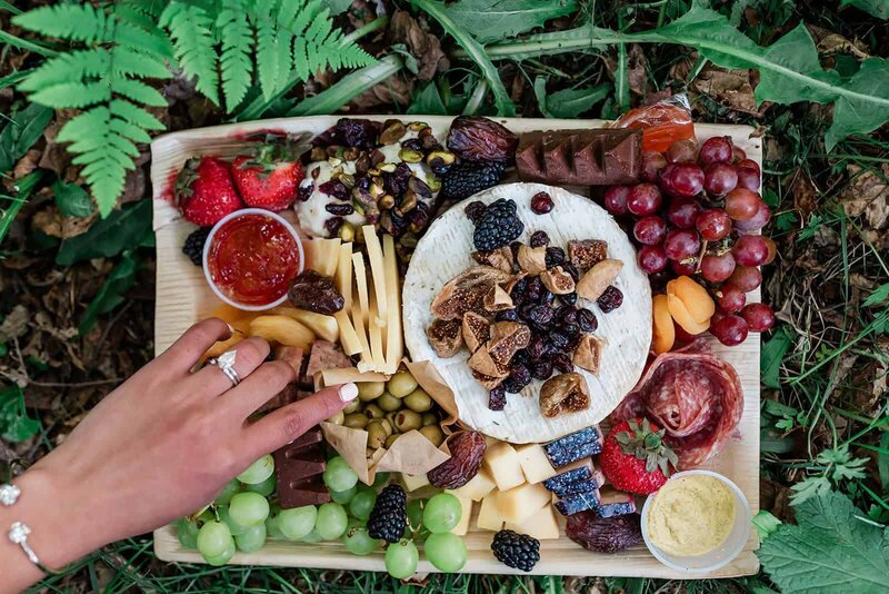 A bride serves herself from a premium charcuterie tray included in their Kenai Fjords adventure luxe elopement package. Her diamond wedding ring catches the light against the lush fern and moss background. 