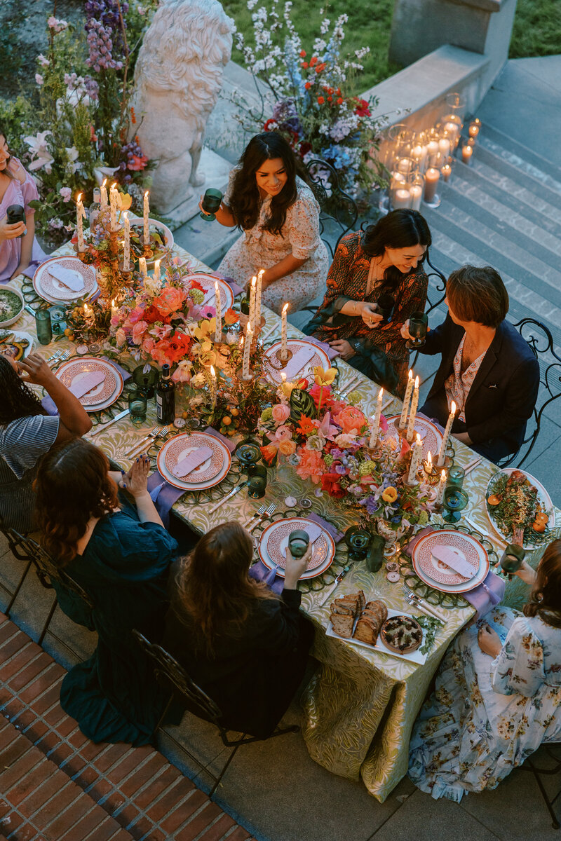 Guests dining alfresco at Wrensmoor Castle. while sitting at a colorful, maximalist, tablescape. Velvet napkins, citron damask linen, taper candles, family-style meal