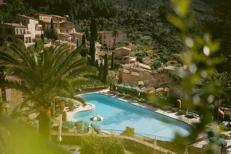 A picturesque hillside village with rustic stone buildings surrounded by lush greenery. A large, clear swimming pool sits in the foreground, bordered by palm trees and lounge chairs, offering a serene retreat under a sunny sky.