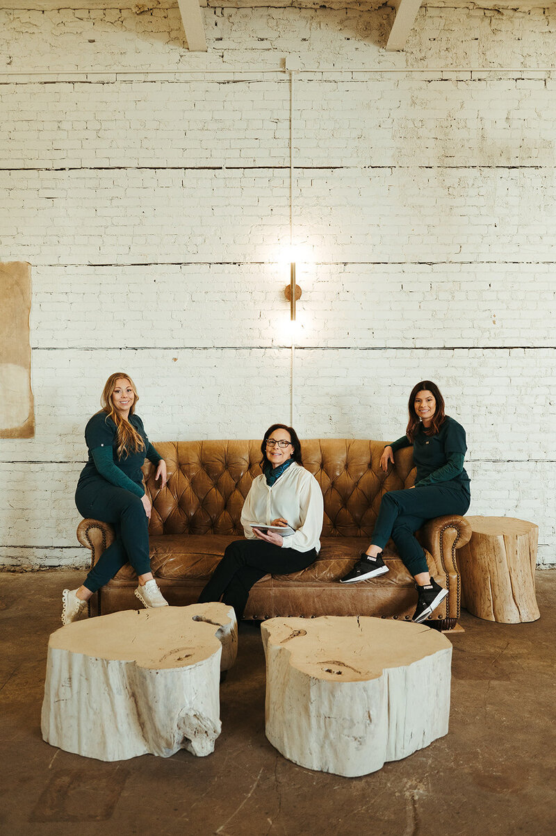 The Kalispell Denture Studio team sits on a leather couch in front of a white brick wall and smiles at the camera.