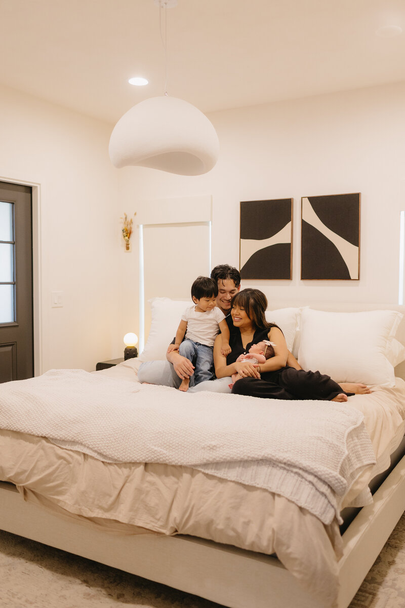 family of four sitting on bed, smiling at each other while holding their newborn during relaxed in-home lifestyle photography session in Monrovia, California