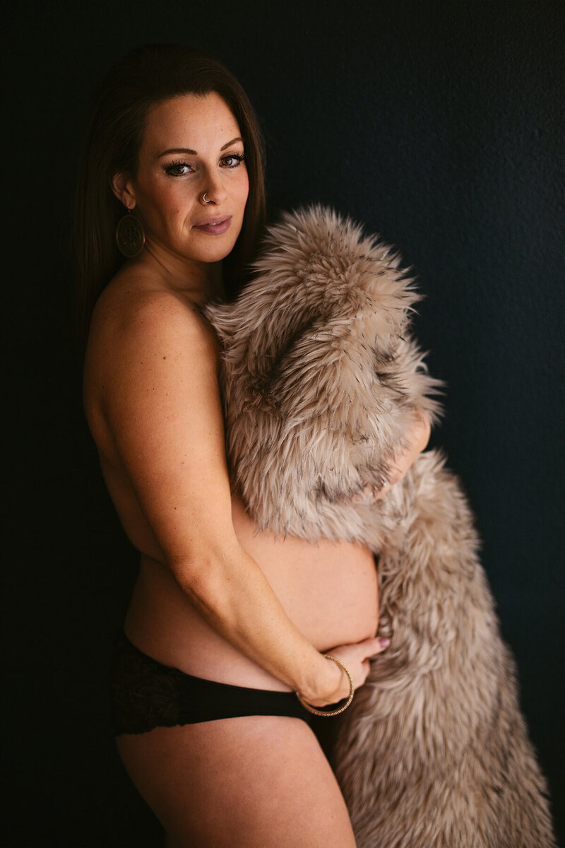 A woman with a large fur blanket covers herself and smiles at the camera at a Bentonville boudoir studio in Northwest Arkansas.