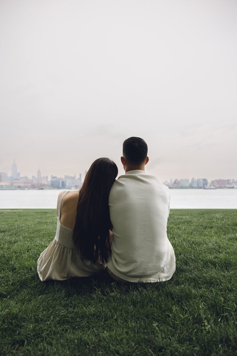 Engaged couple sitting together by the water overlooking Philadelphia skyline during candid engagement session