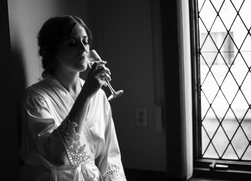 Bride in white satin and lace robe sits in a picture window sipping a glass of champagne