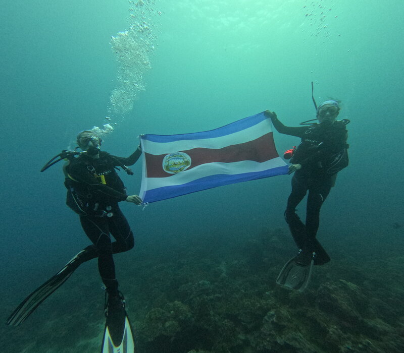 Two female scuba divers hold up the Costa Rican flag mid dive above some coral and rock formations