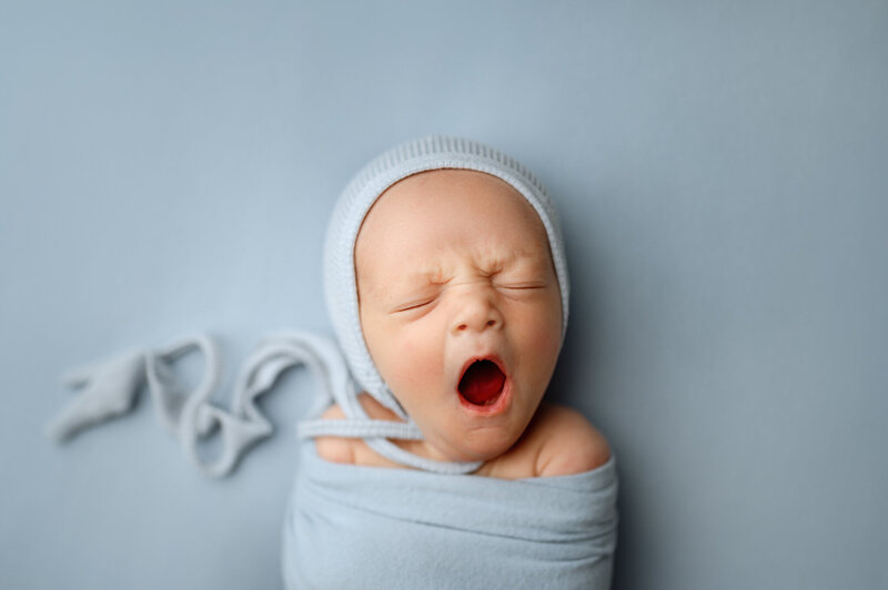 Newborn baby wrapped in soft blue fabric, yawning while wearing a matching knit bonnet. Captured by Honeywild Photo, a Grand Rapids newborn photographer specializing in artistic and timeless baby portraits