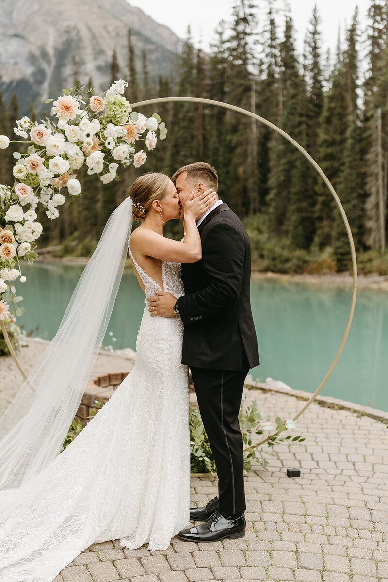 Bride and groom kissing beneath pastel floral arch with mountain backdrop at Emerald Lake Lodge, planned and styled by Melissa Alison Events.