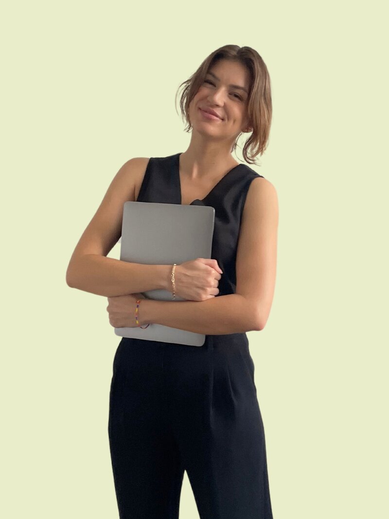Caitlin stands holding a laptop with a relaxed smile. She wears a black vest and black trousers against a green background.