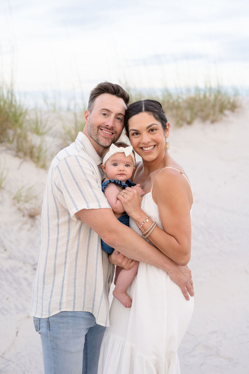 father. mother and baby girl embrace one another while standing on the beach with the dunes, grasses, and ocean in the background