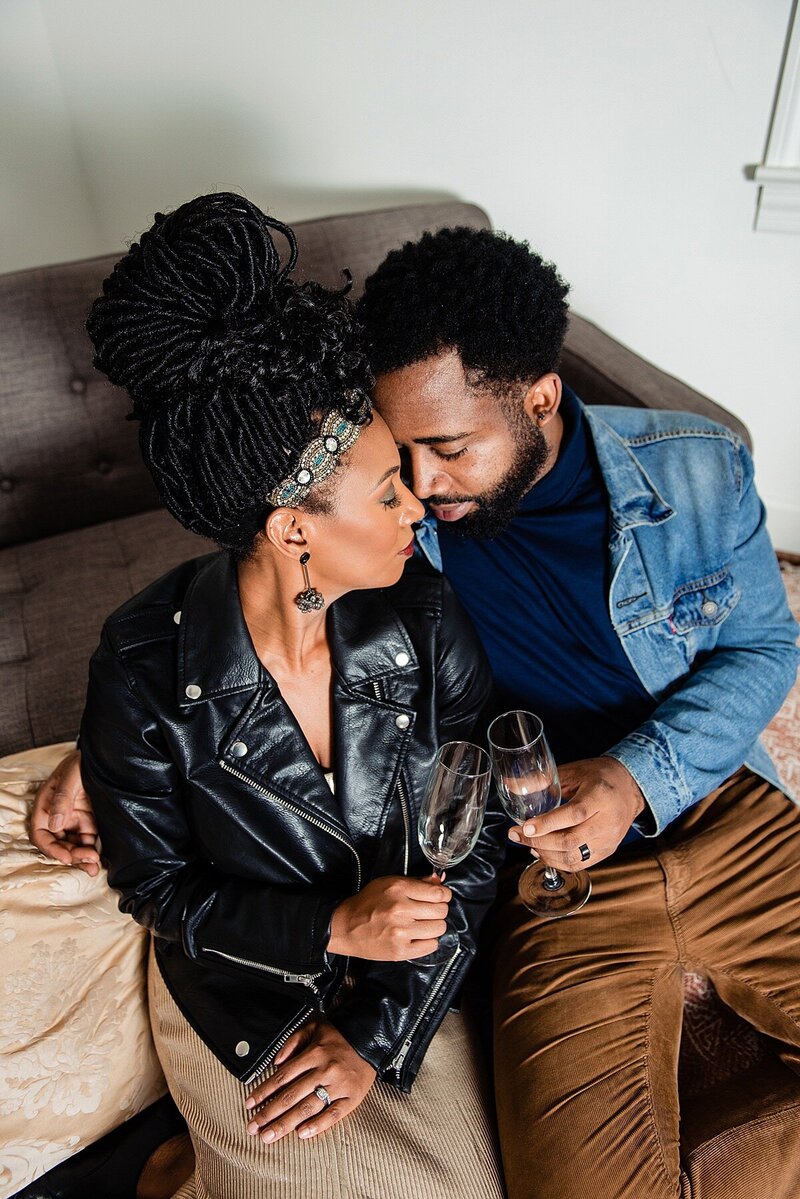 Couple sitting on a couch holding glasses of champagne and sharing a quiet moment together, foreheads leaned together