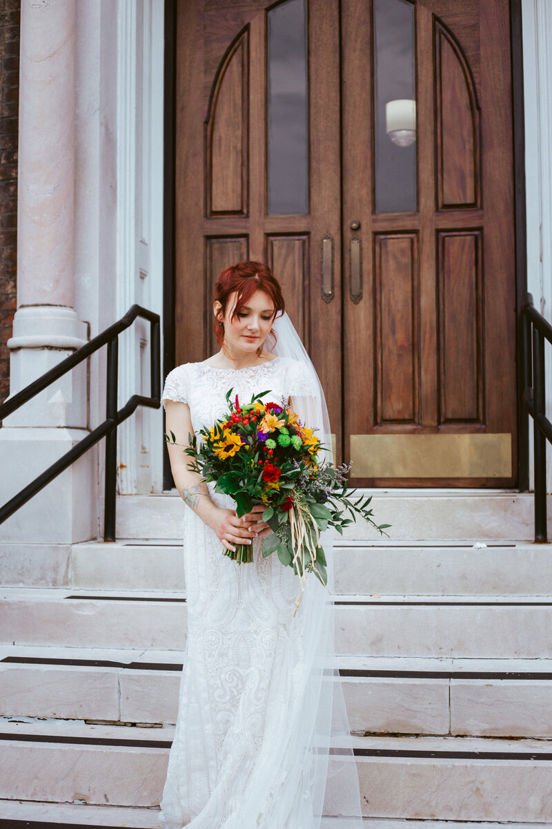 bride with red hair in white dress holding colorful flowers on church steps