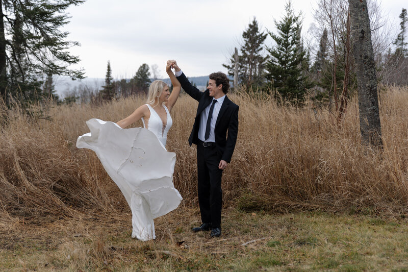 Adventurous elopement photographer captured groom twirling bride in nature, her dress flowing in the breeze with water in the background.