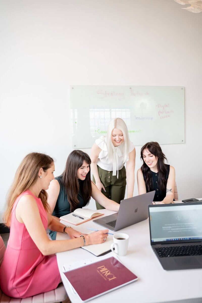 Four Pierce PR team members collaborate at a table, smiling and discussing a project on a laptop.
