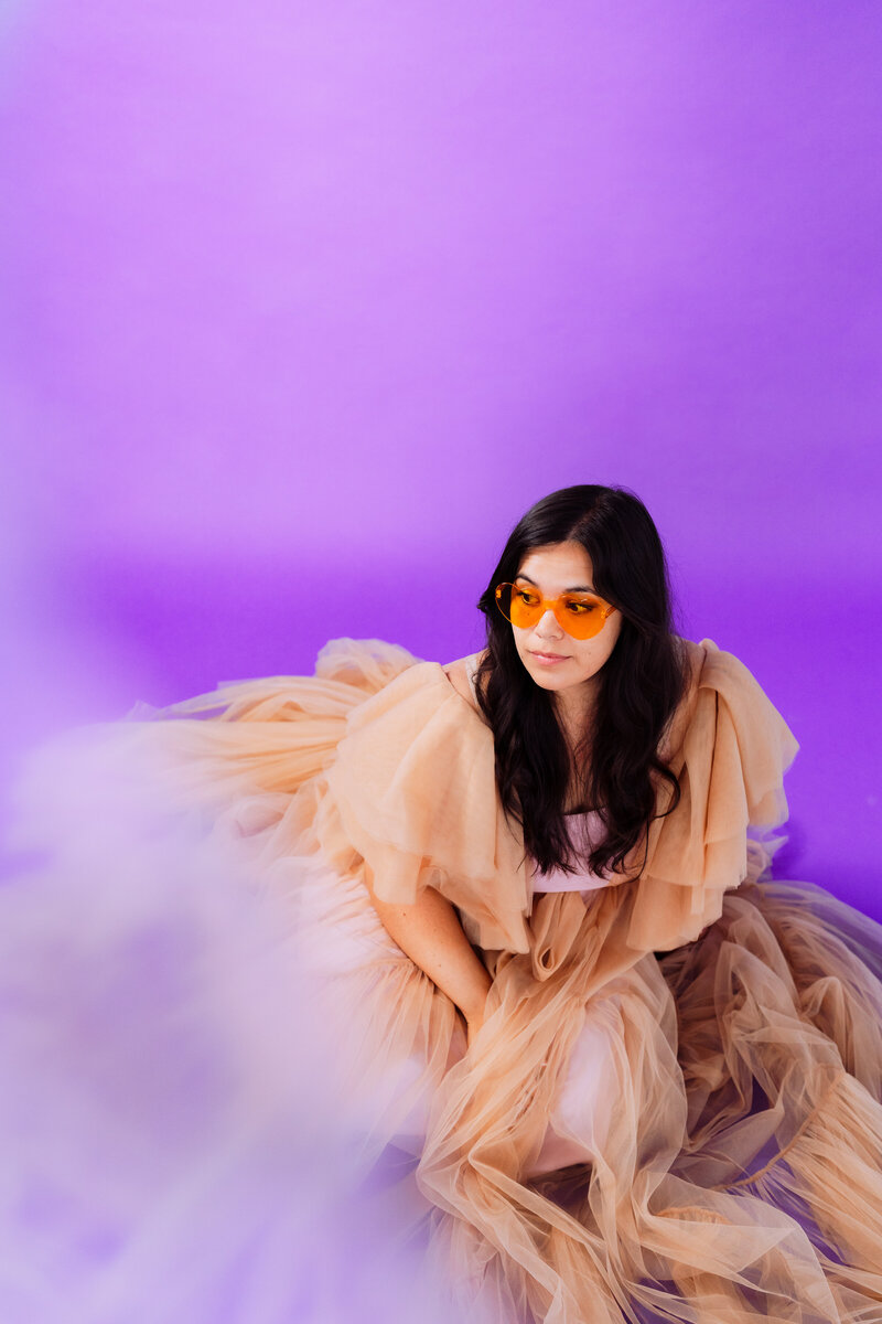 Artistic brand portrait of creative woman in flowing tulle gown and orange sunglasses against a vibrant purple studio backdrop – high-impact studio session by Mewphotovideo