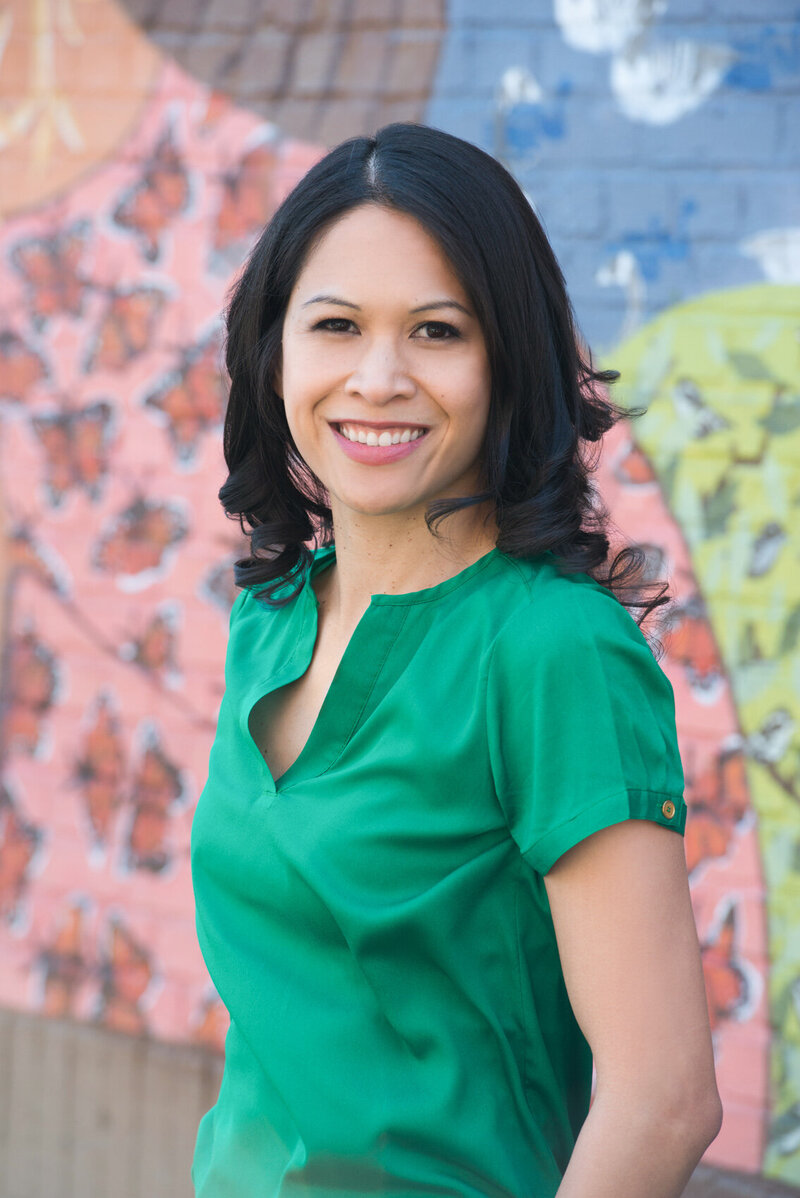Dr. Jen, a Filipino woman with shoulder-length dark hair, leans against a colorful mural painted on a brick wall behind her, smiling at the camera.
