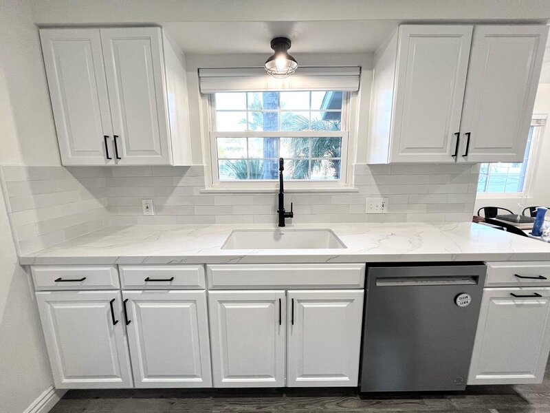 A remodeled kitchen with new white cabinetry and black hardware. There is a fresh white backsplash and white countertops. The sink overlooks a window and has a black light above.