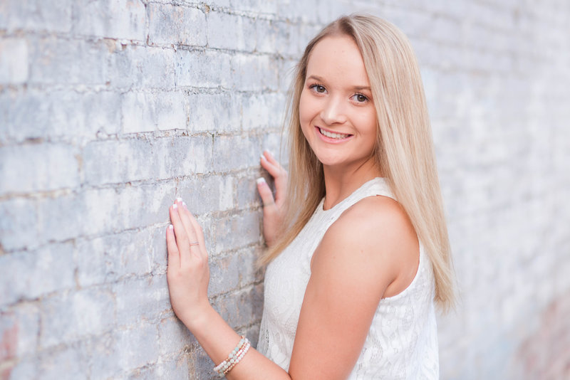 triway high school senior girl facing a powder blue brick wall with hands on wall looking over left shoulder towards camera smiling photographed in wooster ohio photographed by jamie lynette photography canton senior photographer