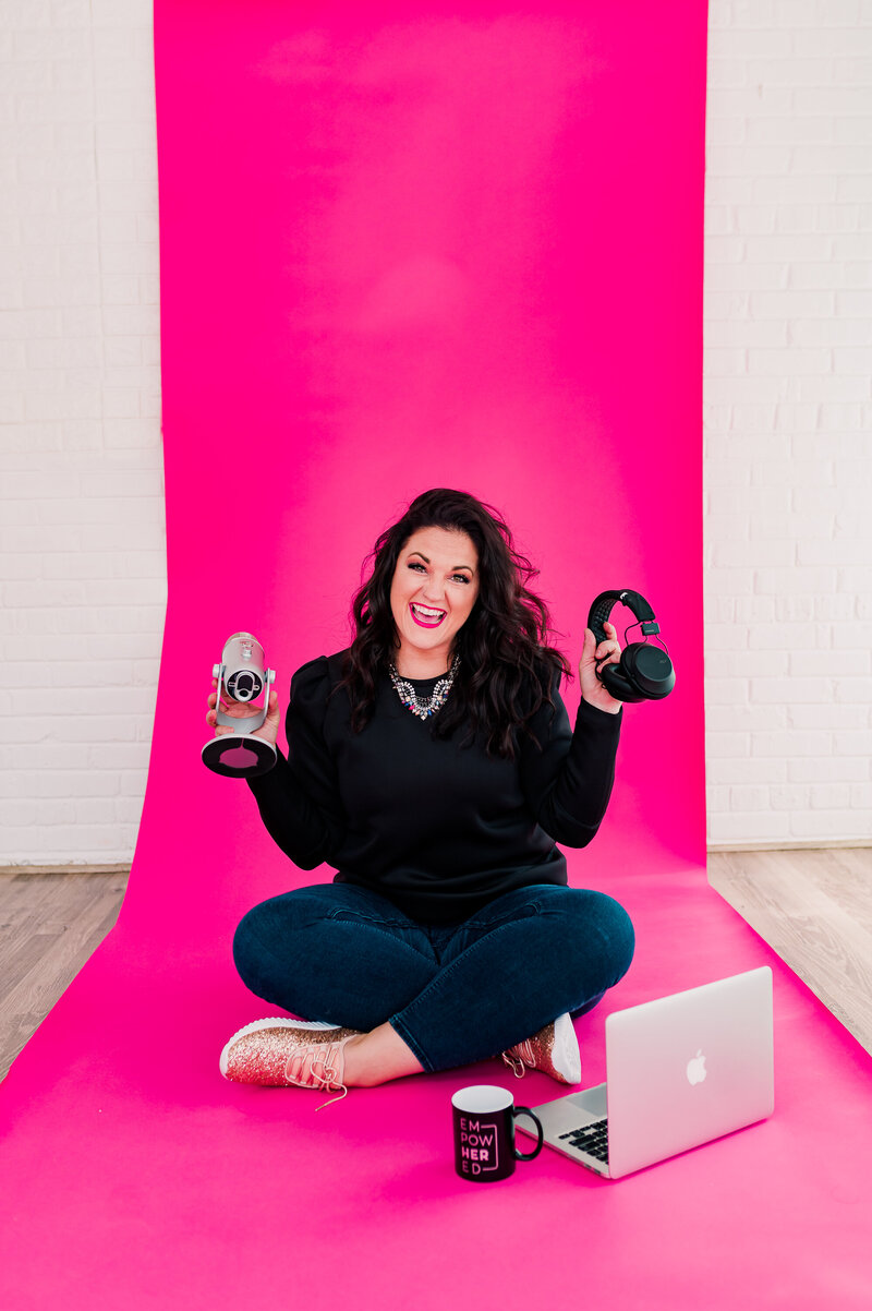 Jessi Cabanin is on a pink background holding a microphone and ear phones with a laptop and coffee cup in front of her.