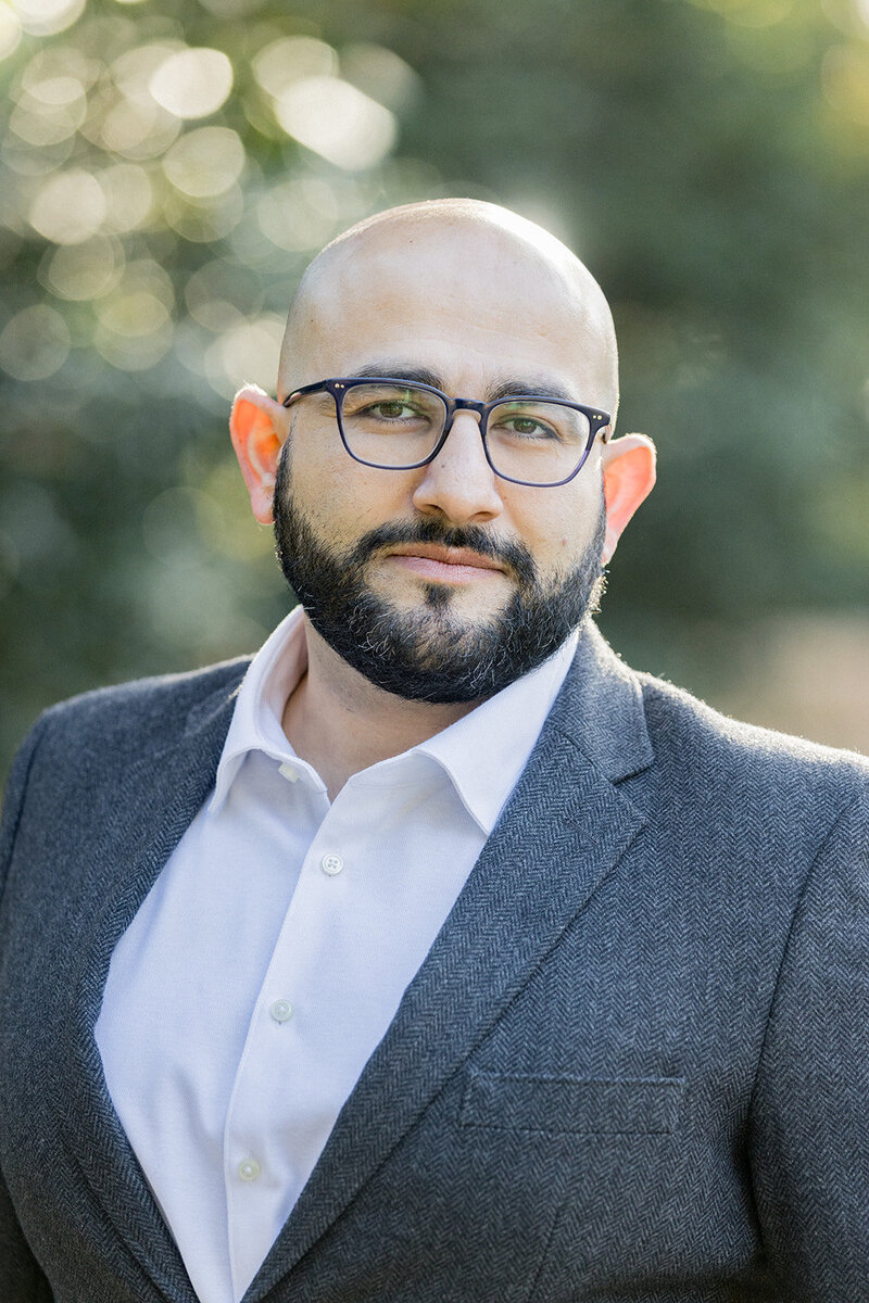 Professional headshot of a confident man in a gray herringbone blazer and white dress shirt, set against a softly blurred outdoor background. Natural lighting highlights his approachable yet authoritative presence. Small business branding photography in Columbia, SC, specializing in corporate headshots and personal branding for professionals.